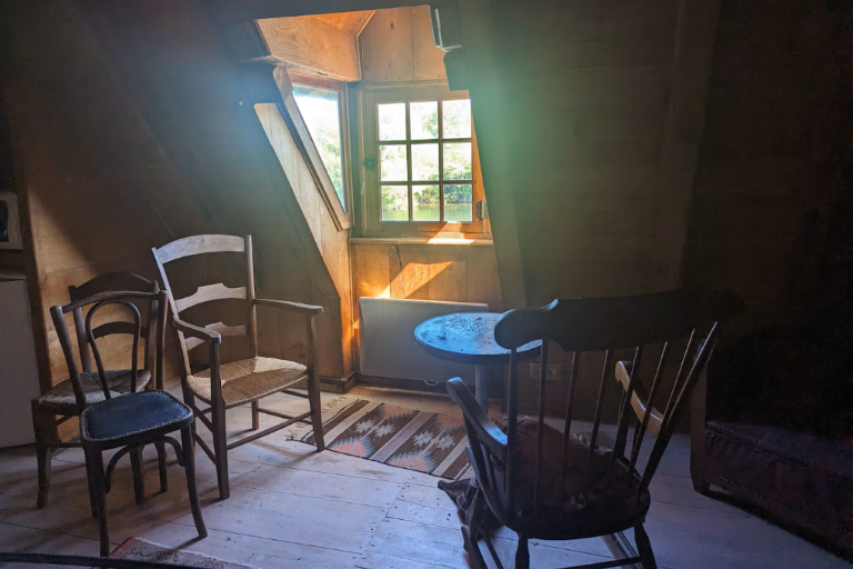 Sun streams through a wood window revealing a cluster of wooden chairs, empty but in slight disarray, suggesting their previous occupants appear to have just left
