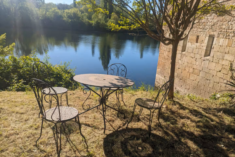 round outdoor table and chairs on a roughly mowed river bank with picturesque view, with the side of the mill visible