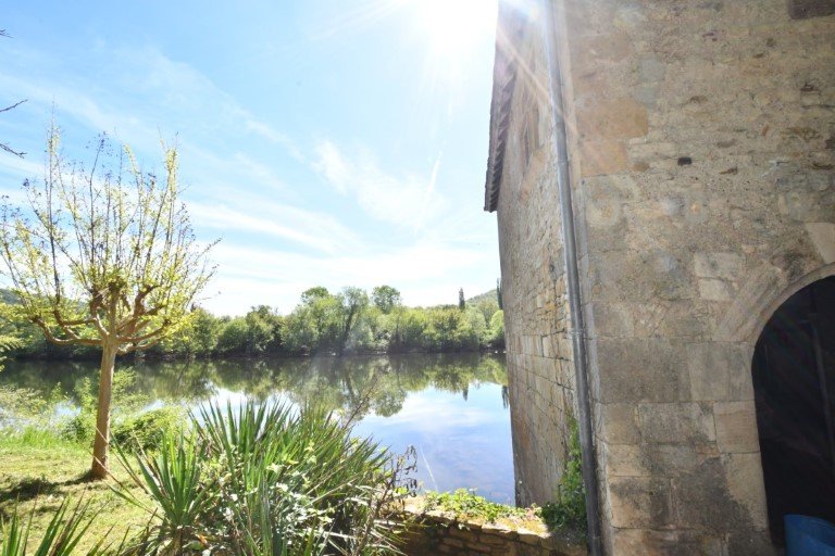 blue sky reflecting on river, viewing across to green trees on other side, with the wall of the mill visible to the right hand side